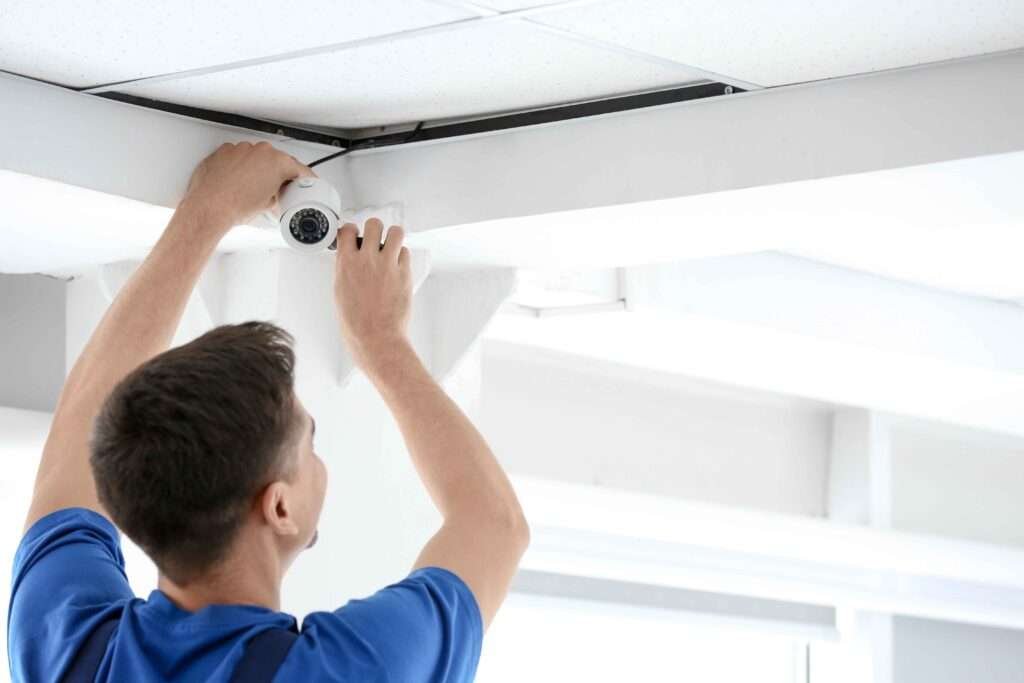 A technician in blue overalls is performing a CCTV installation, mounting a white dome security camera onto a ceiling beam in a commercial building